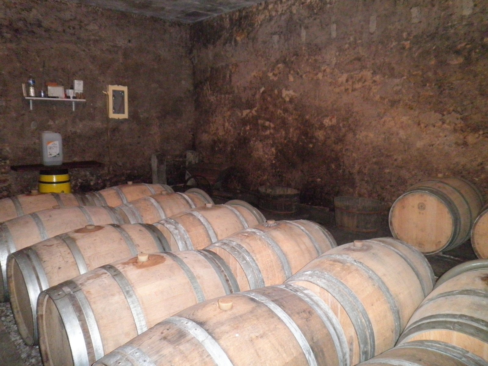 Des rangées de barriques en chêne sont couchées sur le sol d'une cave aux murs de pierre brute, avec une étagère et un bidon au fond.