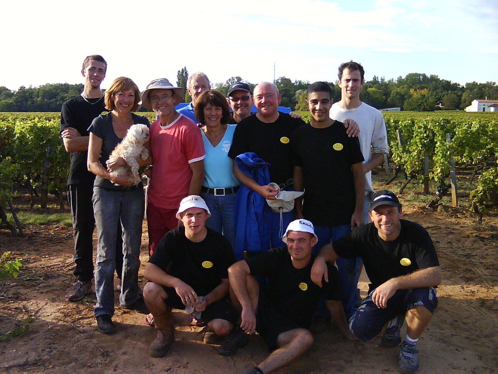 Un groupe de onze personnes souriantes, dont certaines accroupies, pose avec un petit chien blanc devant des rangs de vigne par une journée ensoleillée.